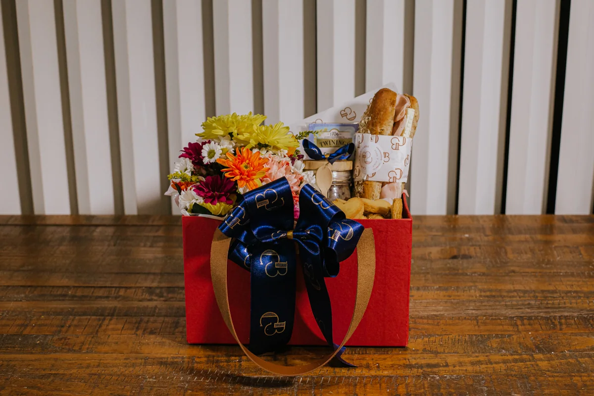 Cesta vermelha com flores, sanduíche e quitutes, decorada com laço azul
